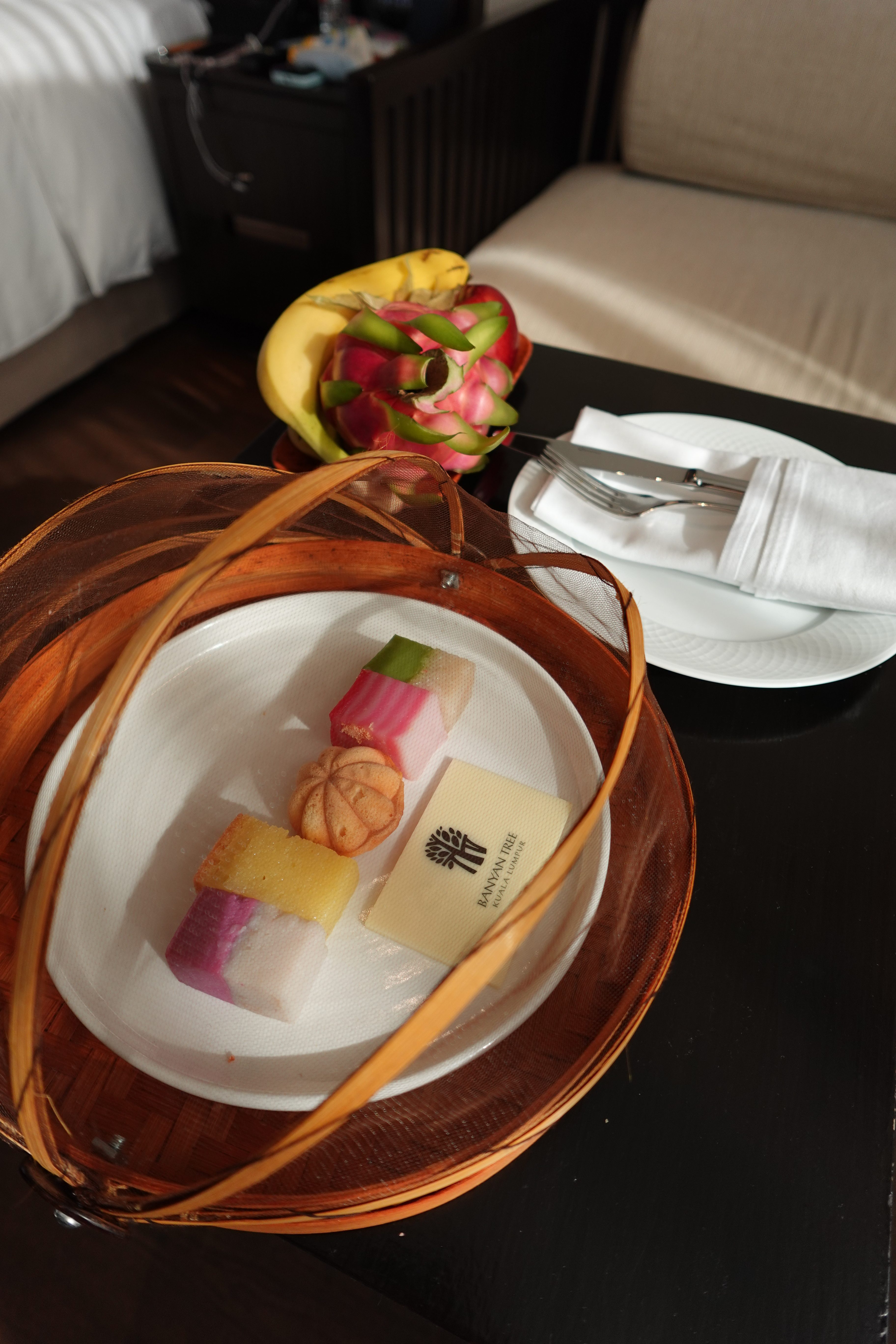A wooden basket containing colorful desserts including layered jelly and a cake, placed on a table with bananas and a dragon fruit in the background.