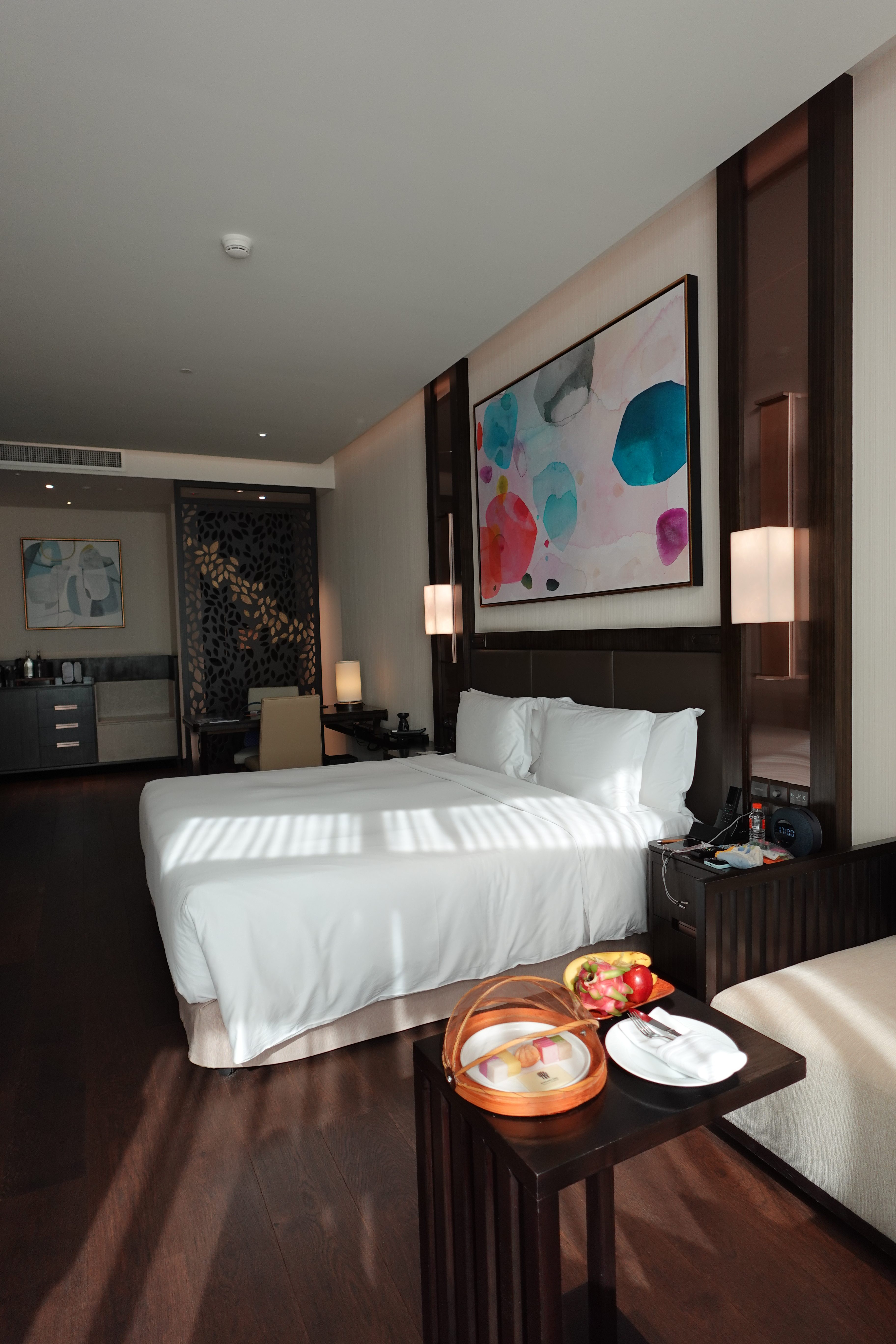 Modern hotel room featuring a queen bed with white linens, a colorful abstract painting on the wall, and a small table with fruits and snacks.