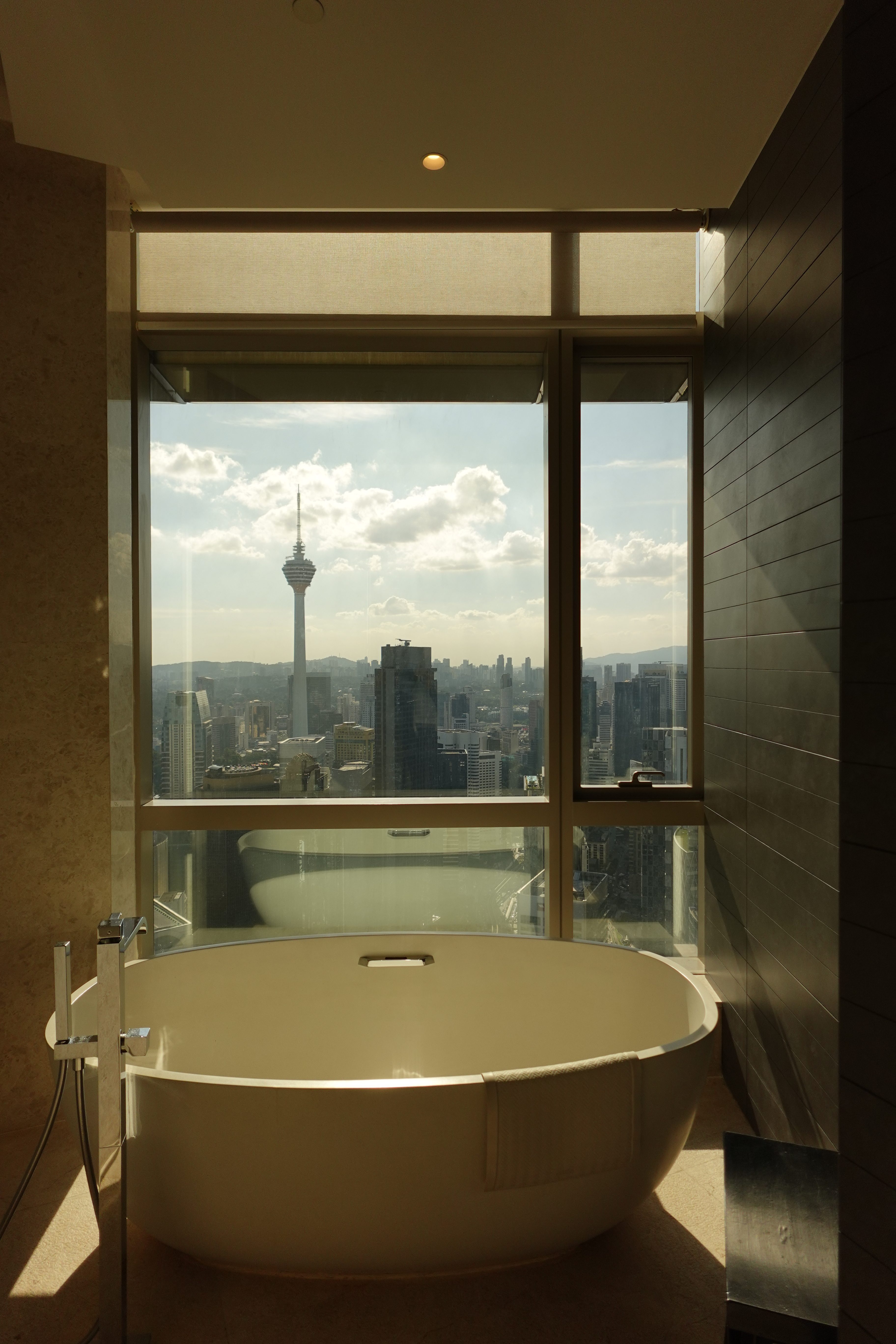 A luxurious bathroom featuring a freestanding bathtub with a panoramic view of a city skyline, including a prominent tower, under a bright sky with scattered clouds.