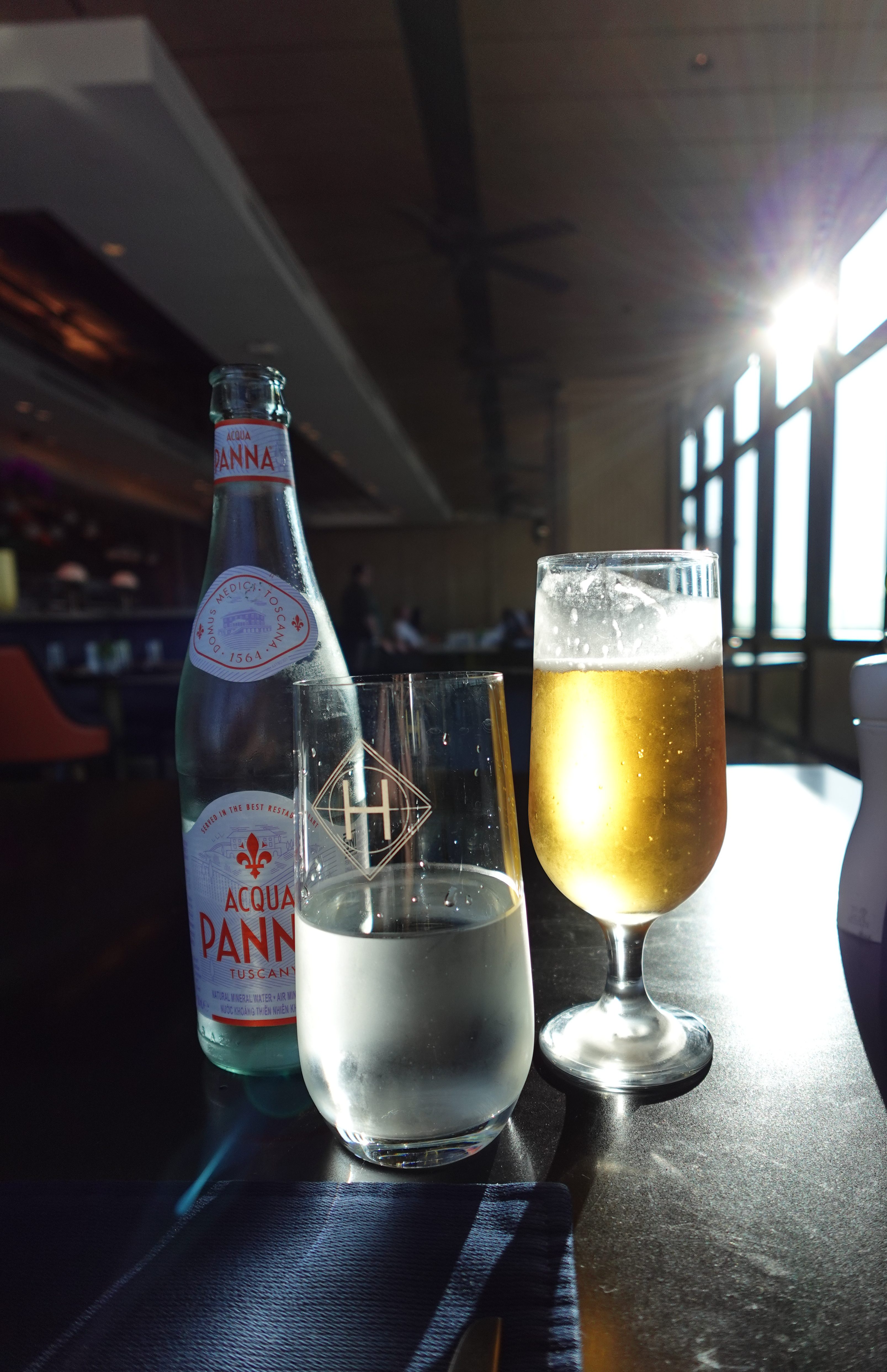 A bottle of Acqua Panna sparkling water next to a glass of clear water and a glass of golden beer on a table, with sunlight coming through windows in the background.