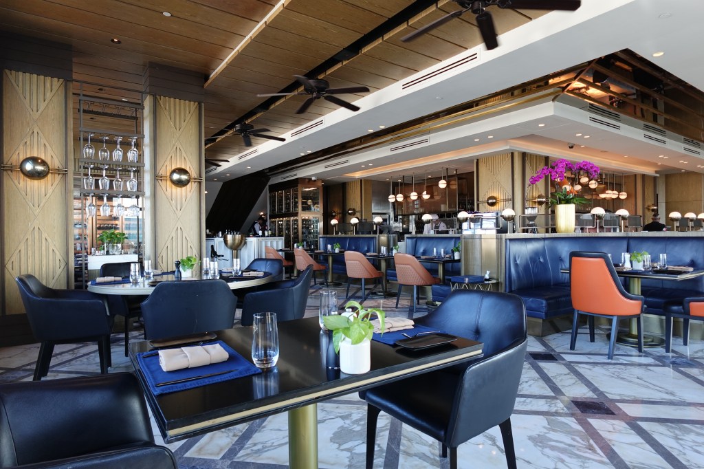 Elegant restaurant interior featuring stylish black chairs, marble flooring, and wooden accents, with tables set for diners and a well-appointed bar area.
