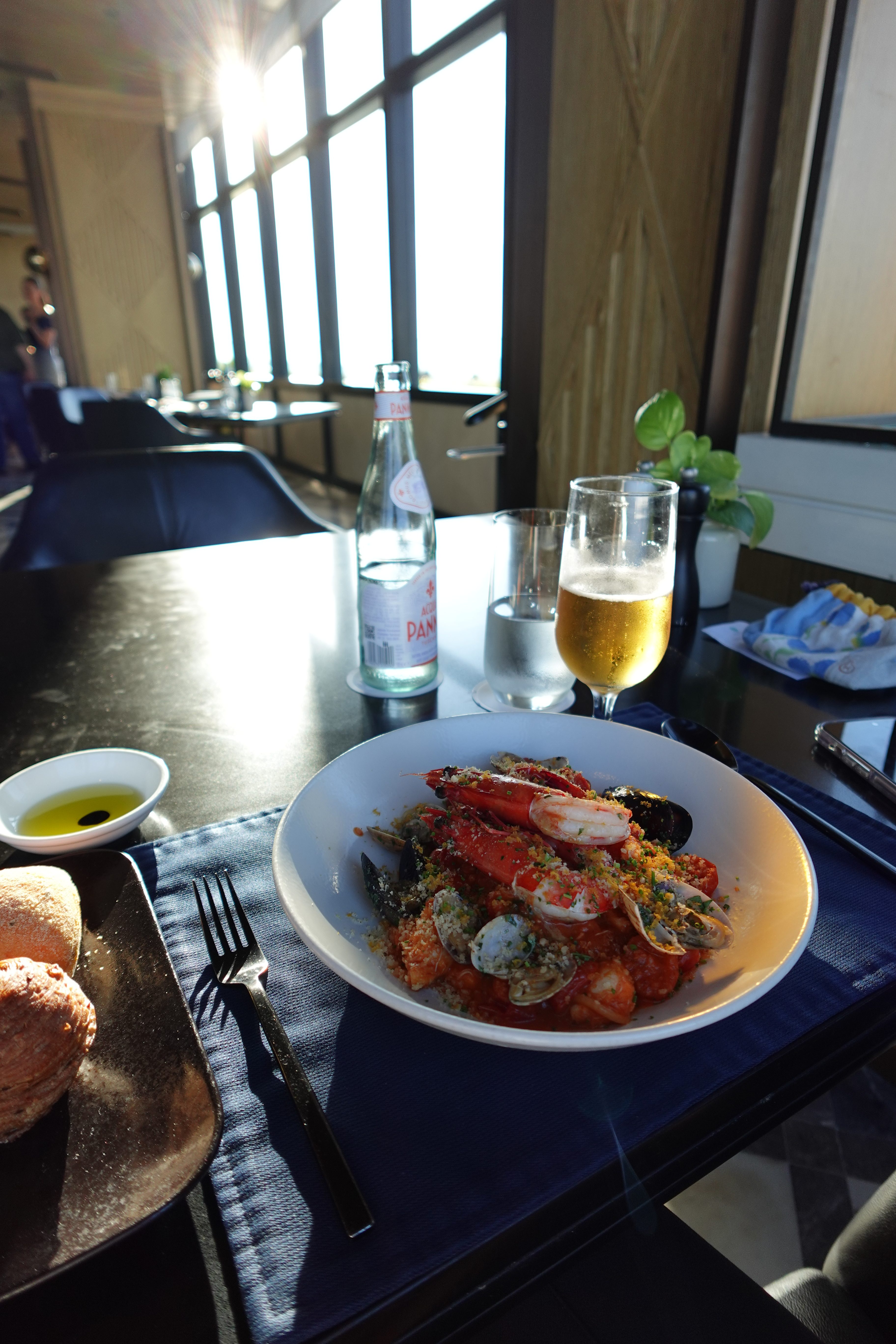 A beautifully presented seafood dish with large shrimp, mussels, and clams on a white plate, accompanied by a glass of beer and sparkling water. The table setting features a small bowl of olive oil, a roll of bread, and elegant tableware, with large windows in the background letting in natural light.