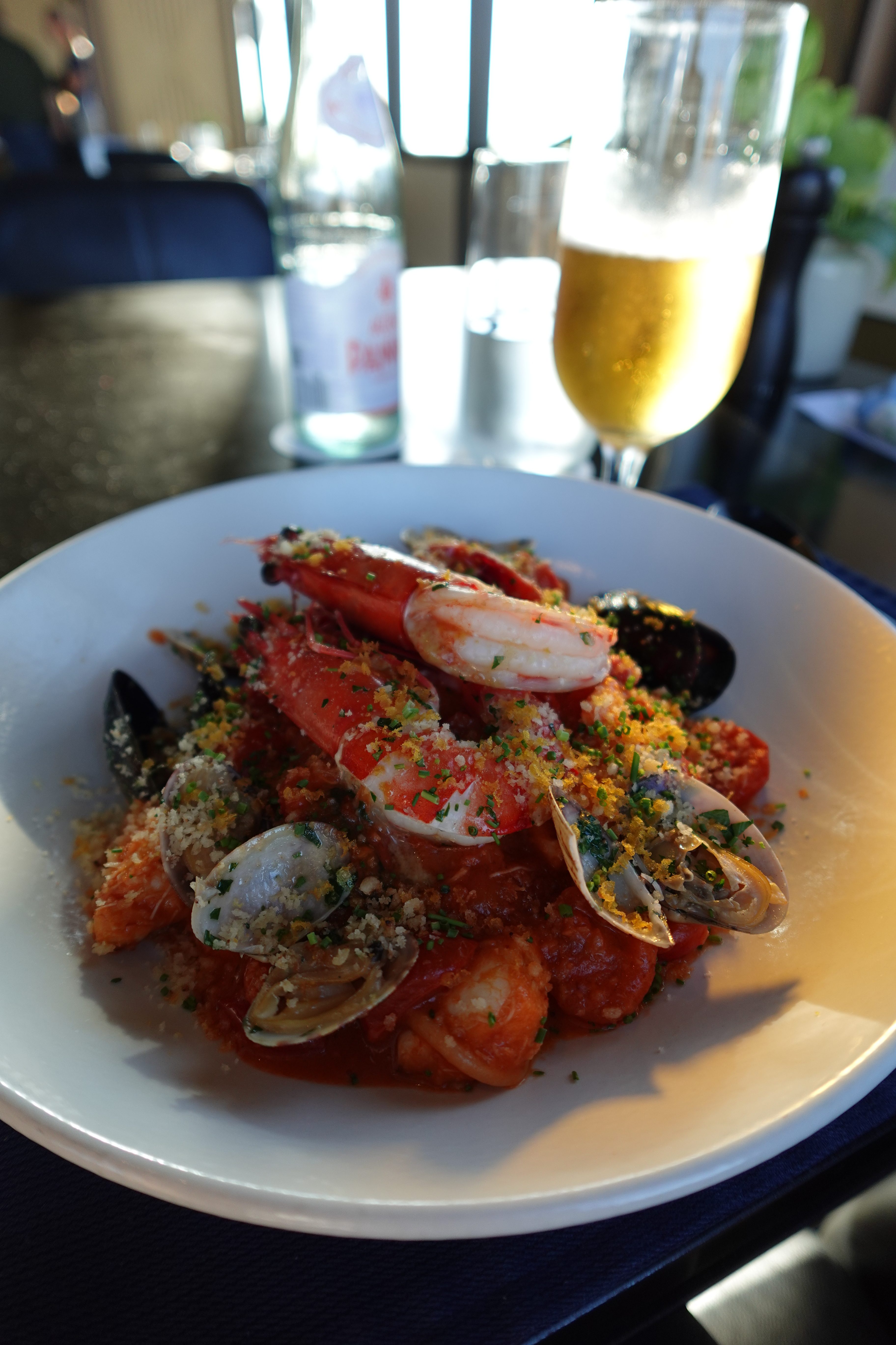 A plate of seafood pasta featuring prawns, mussels, and clams, topped with breadcrumbs and herbs, alongside a glass of beer and sparkling water in the background.