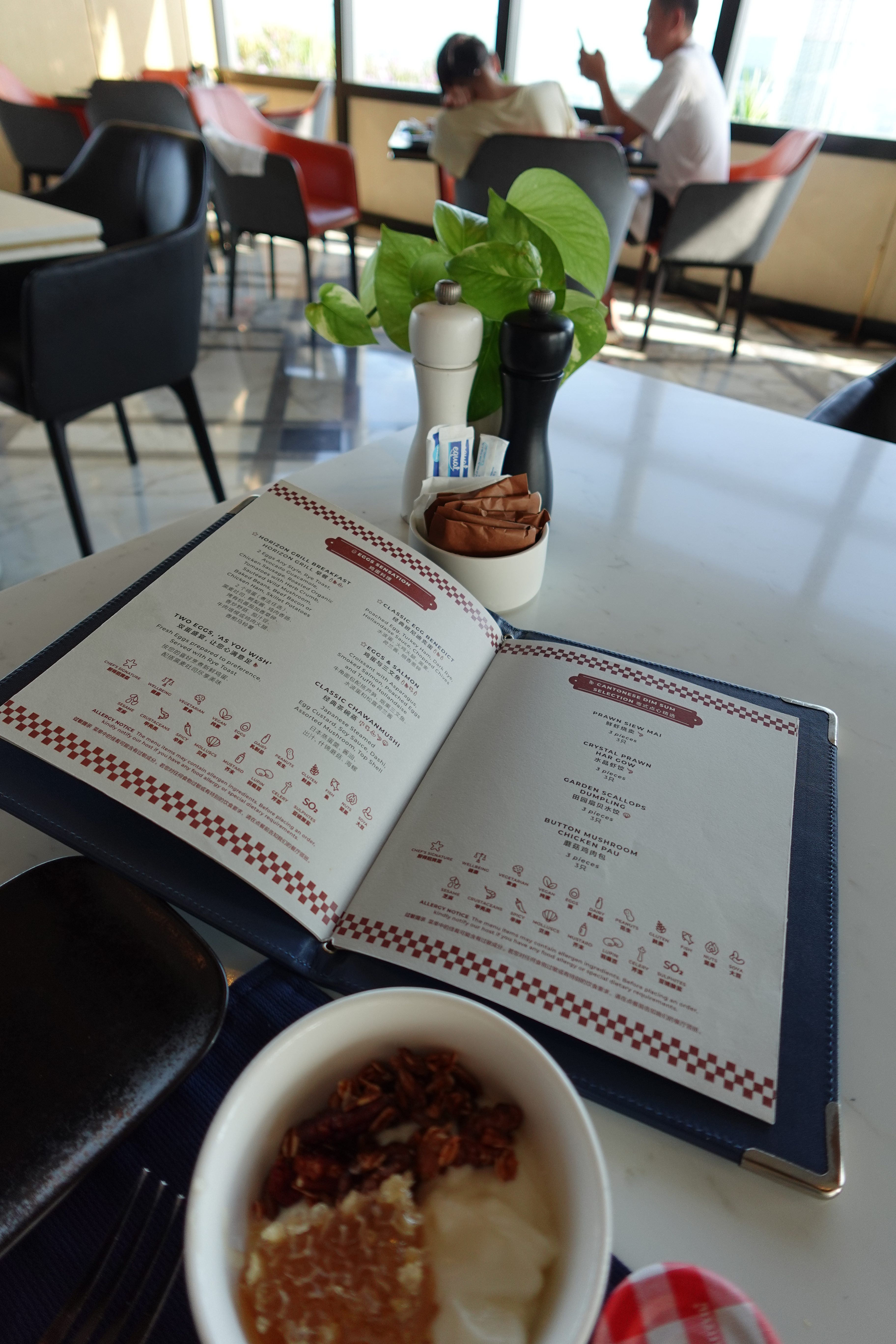 A table setting in a restaurant with an open menu, a bowl of yogurt topped with granola and honey, and a decorative plant. In the background, there's a couple seated at a table.