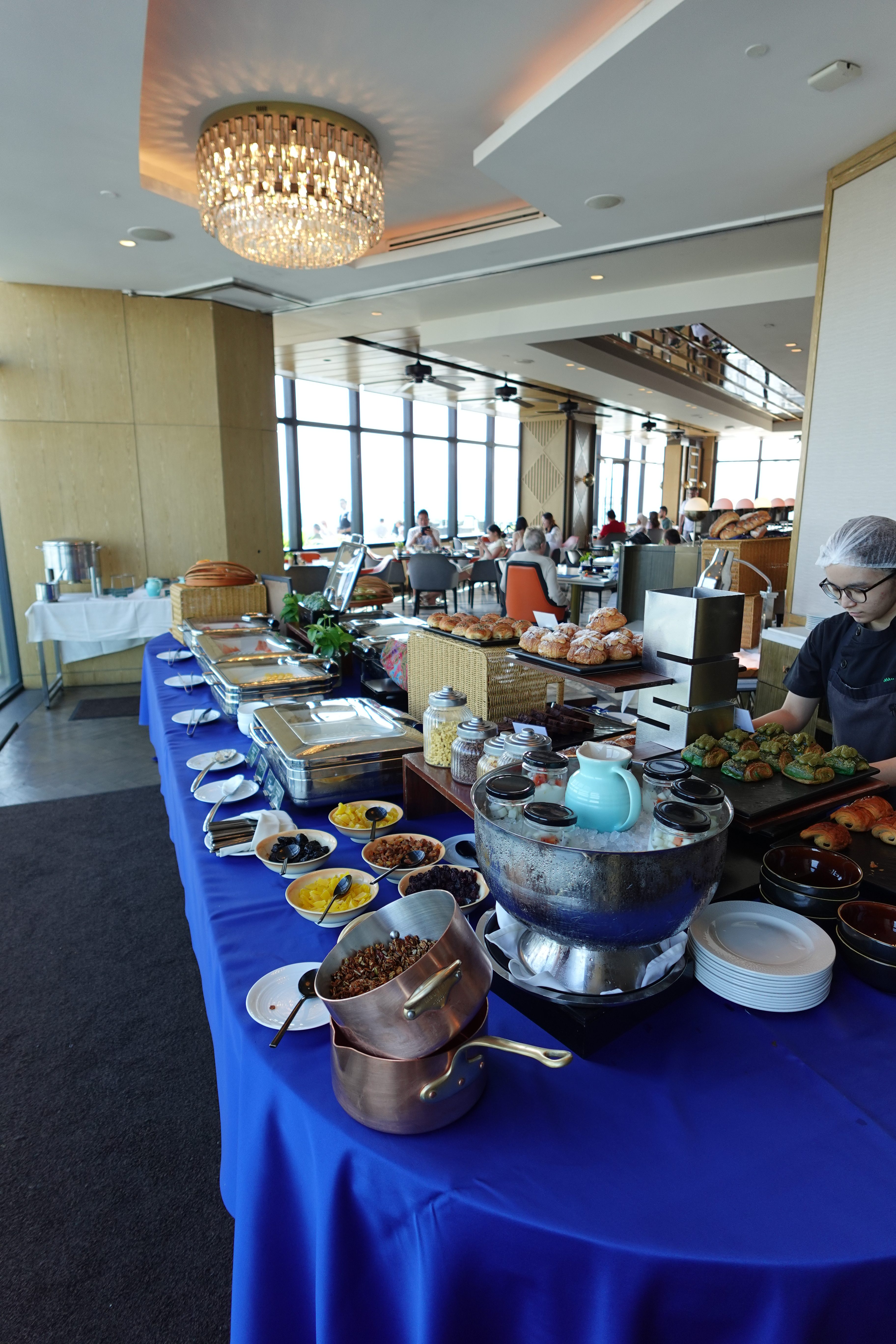 A buffet setup featuring various dishes on a blue tablecloth in a modern restaurant environment, with large windows, vibrant decor, and guests dining in the background.