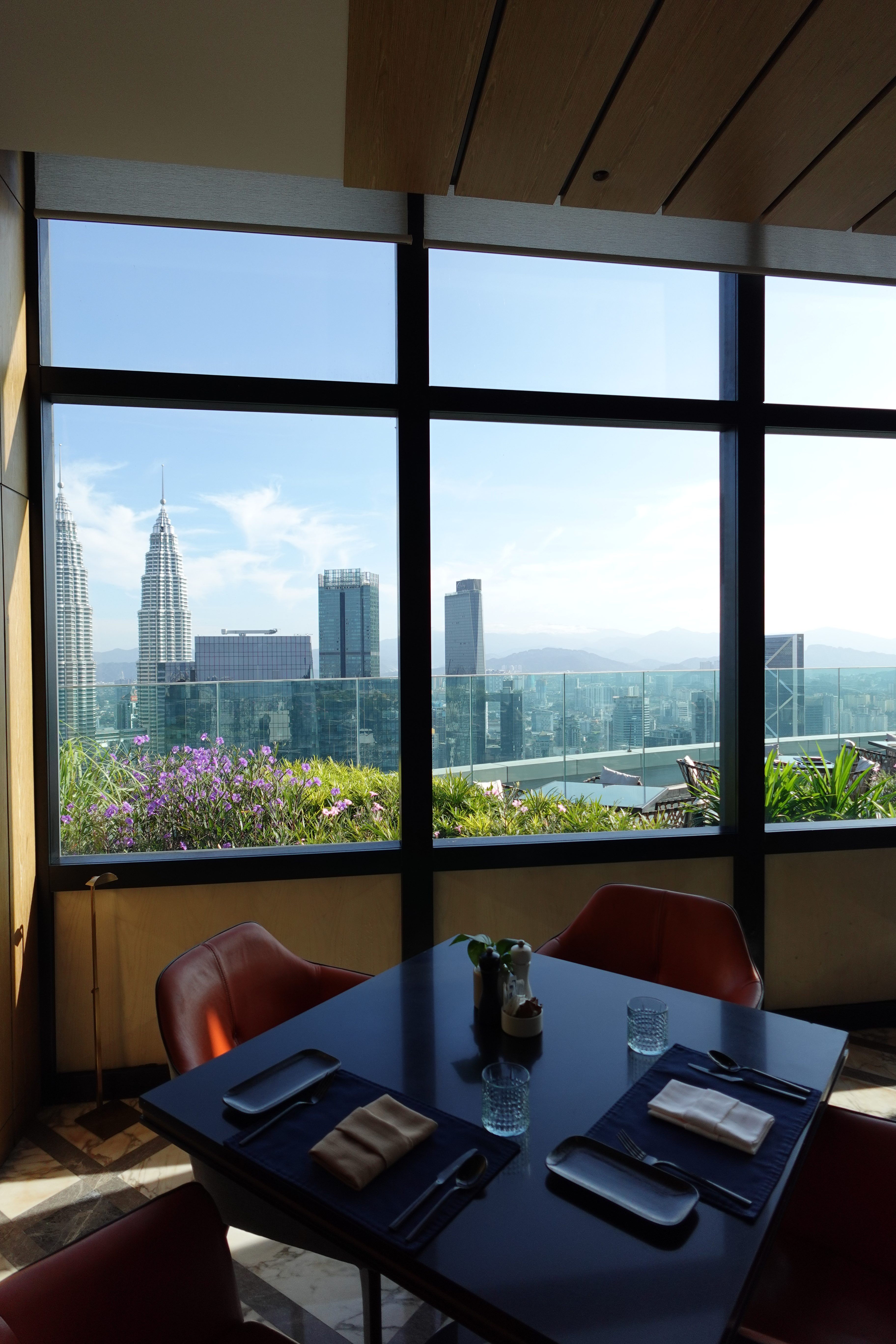 Interior view of a restaurant with a glass window showcasing a city skyline, featuring the Petronas Towers and modern buildings in the background, along with a table set for dining.